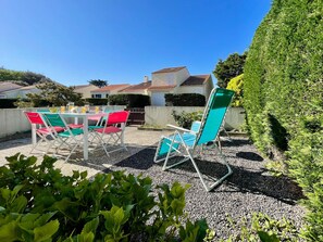 En udendørs terrasse med bord og stole, en hæk og et hus i baggrunden.