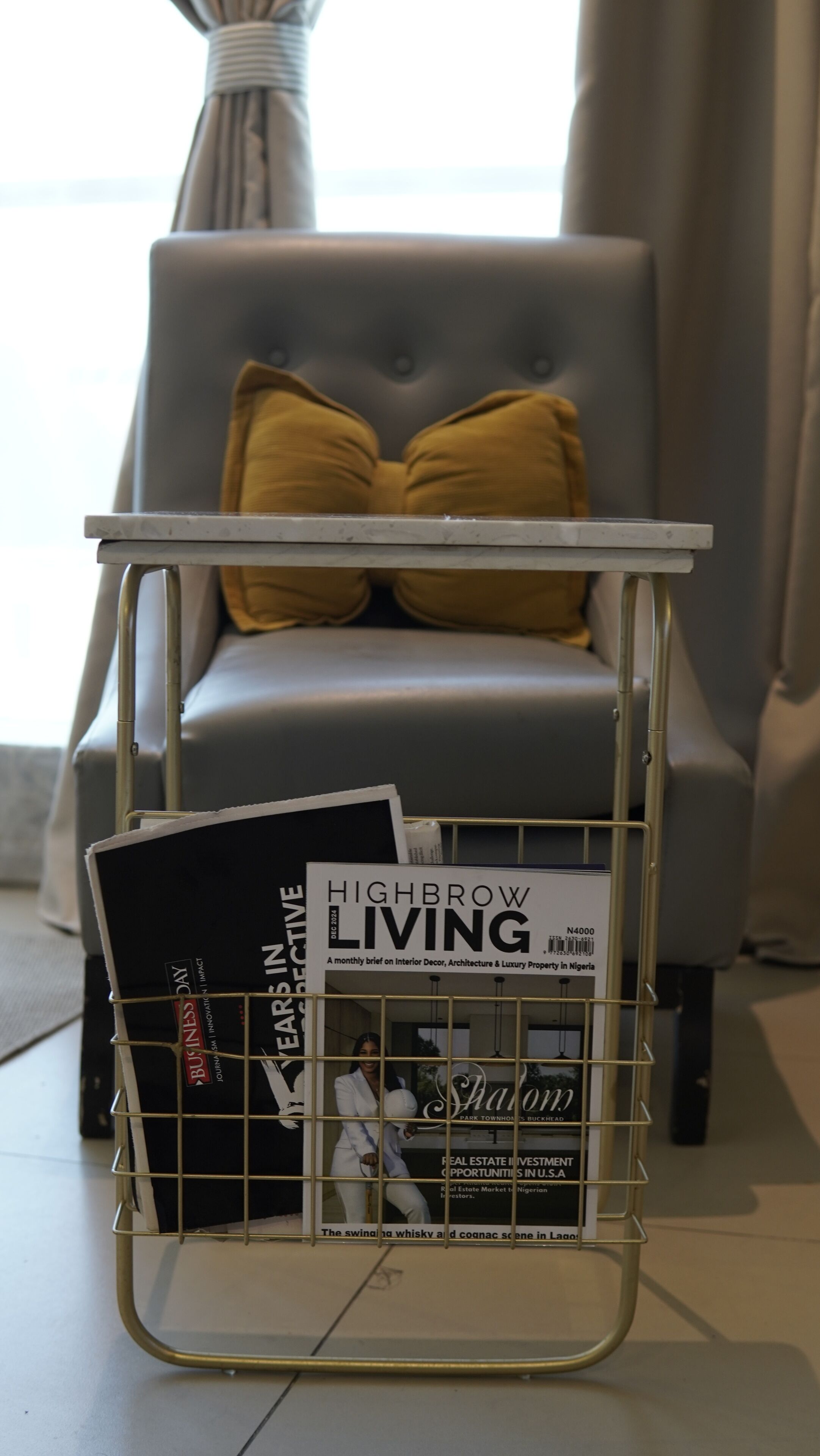 Ein Zeitschriftenständer mit den Zeitschriften 'Highbrow Living' und 'Business Day', platziert vor einem grauen Sessel mit einem gelben Kissen.