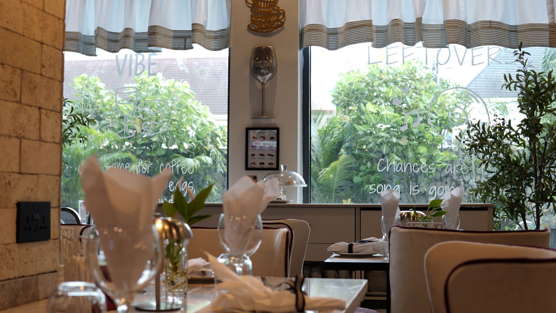 A restaurant interior with tables set for dining, a window with a view of greenery, and a menu board.