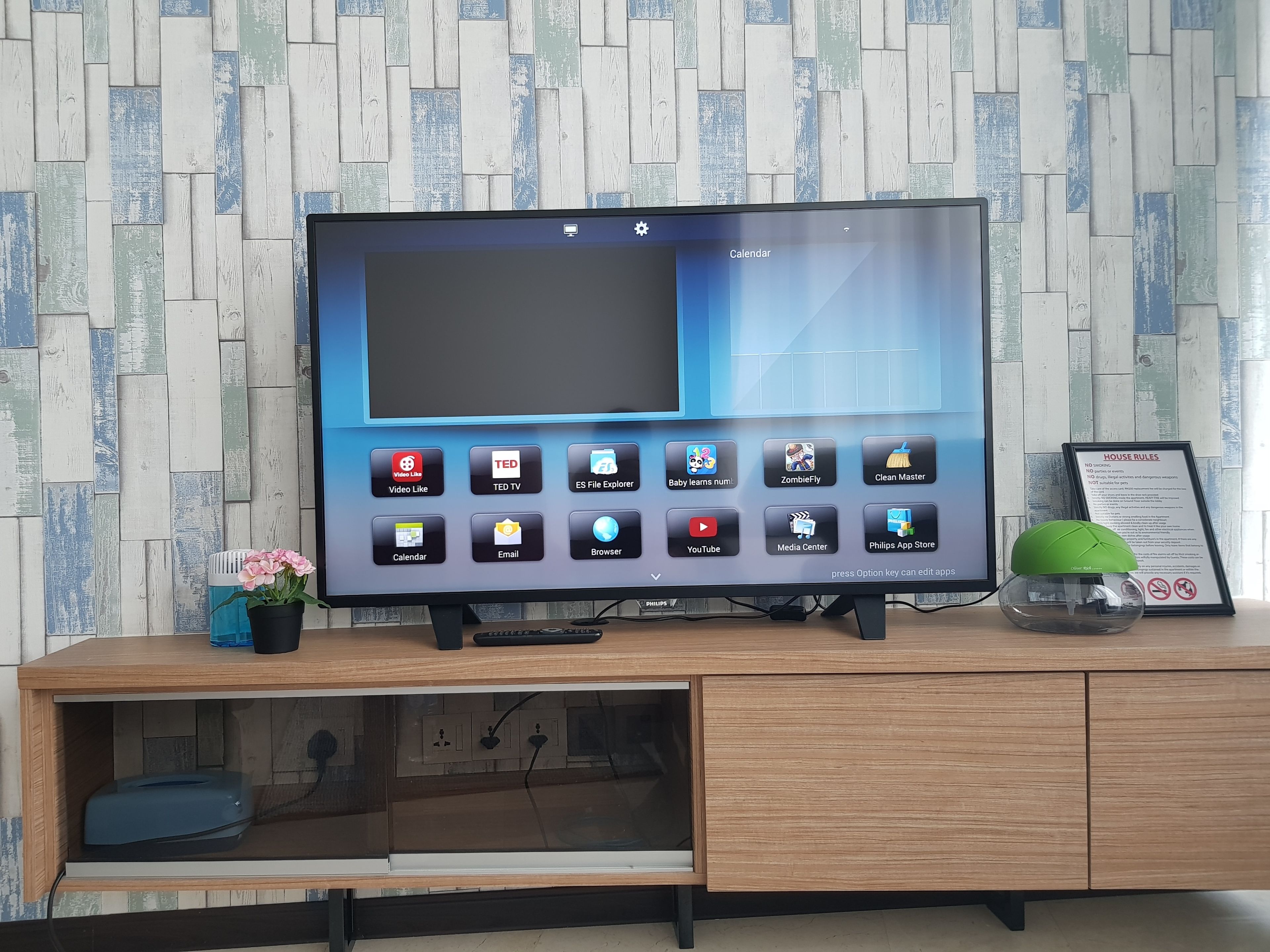 A television with a variety of apps on its screen, placed on a wooden cabinet with a wall-mounted shelf below.