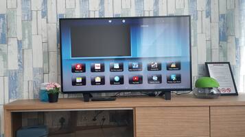 A television with a variety of apps on its screen, placed on a wooden cabinet with a wall-mounted shelf below.