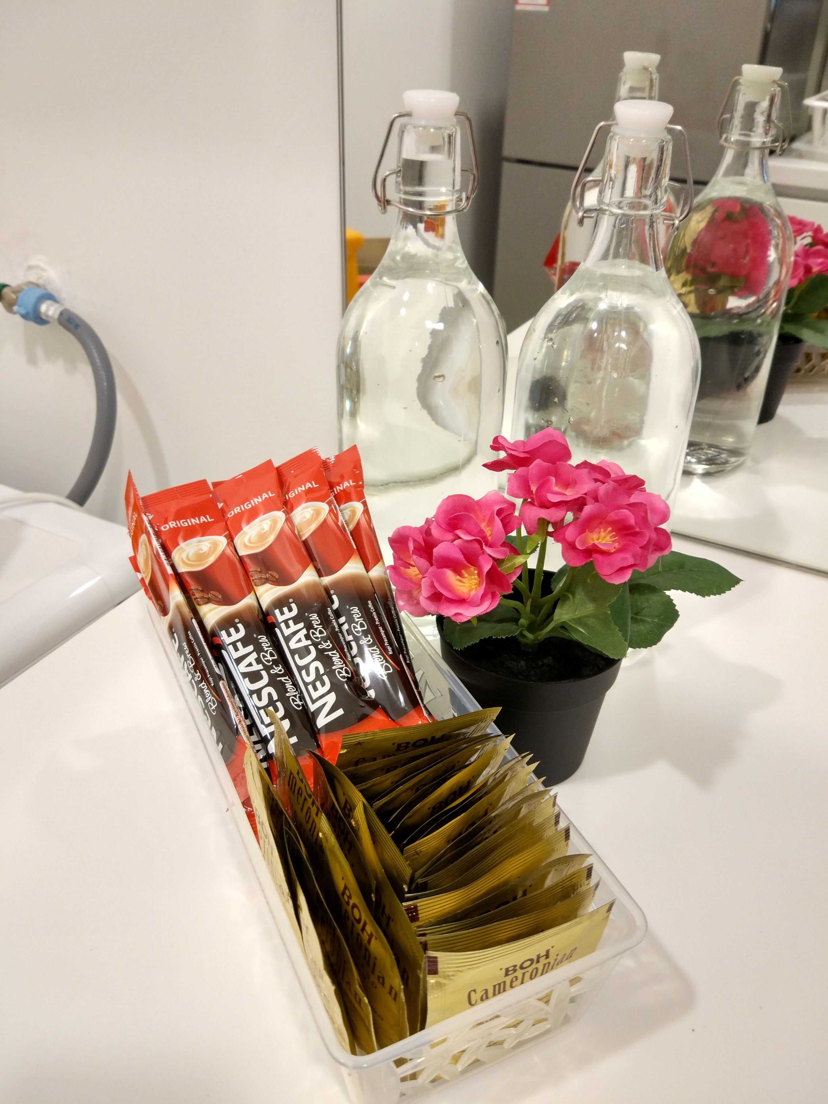 Una bandeja con sobres de Nescafé, una planta en maceta con flores rosadas y varias botellas vacías sobre una encimera blanca.