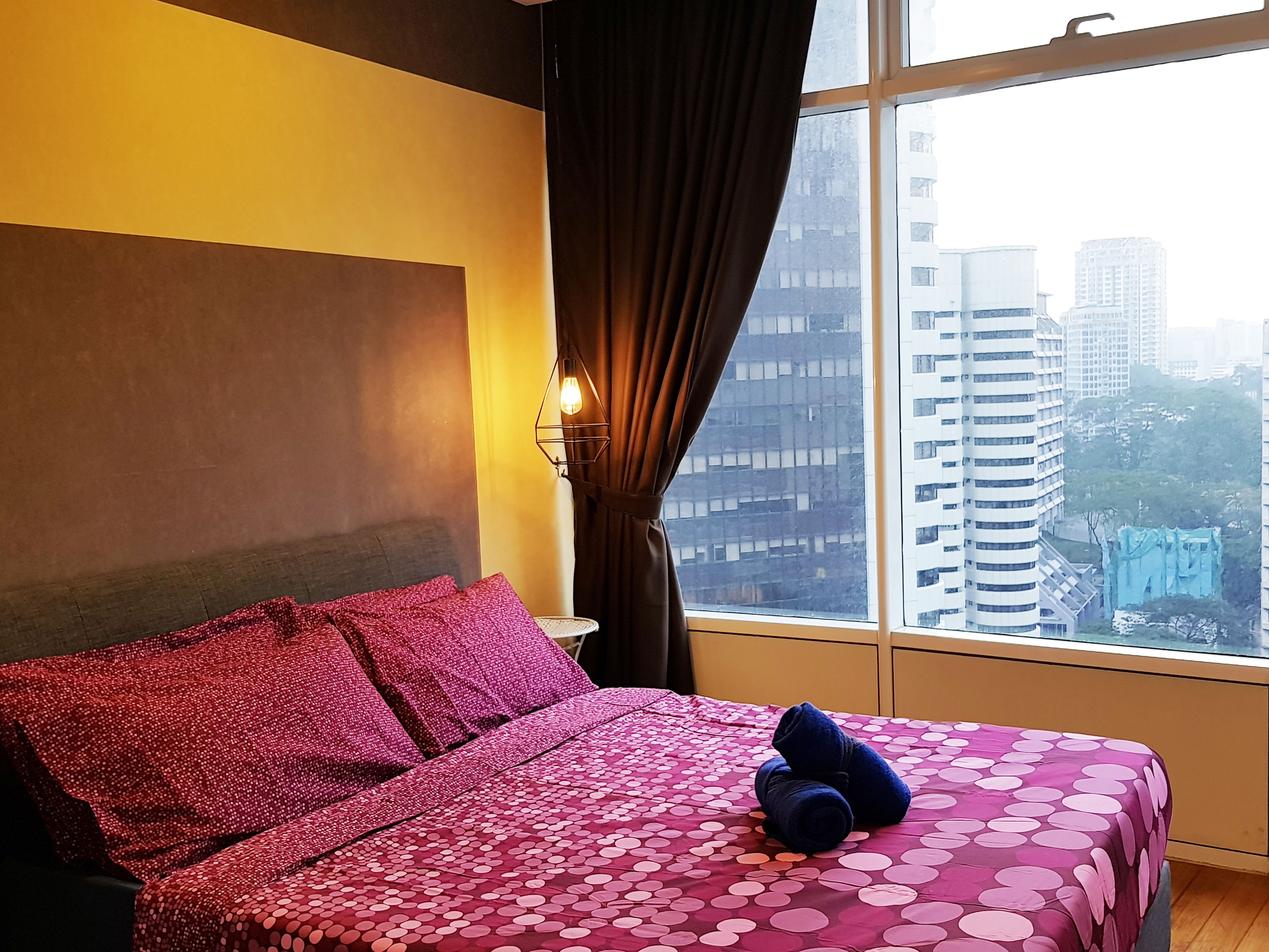 Ein Hotelzimmer mit Bett, Nachttisch, Lampe und Fenster mit Blick auf die Stadt.