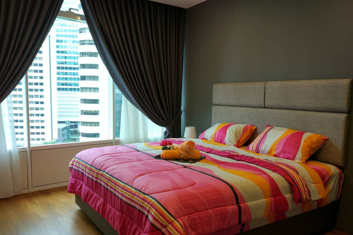 A hotel room with a large bed, grey headboard, and a view of tall buildings through the window.