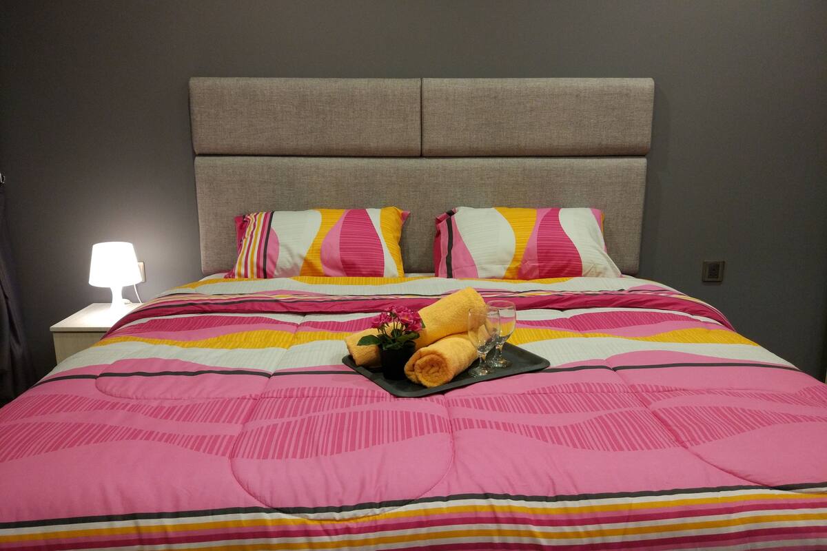 A neatly made bed with striped bedding and pillows, a bedside lamp, and a tray with towels and glasses.