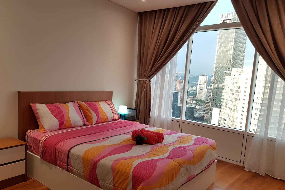 A modern bedroom with a large bed, a nightstand, and a view of a cityscape through large windows.