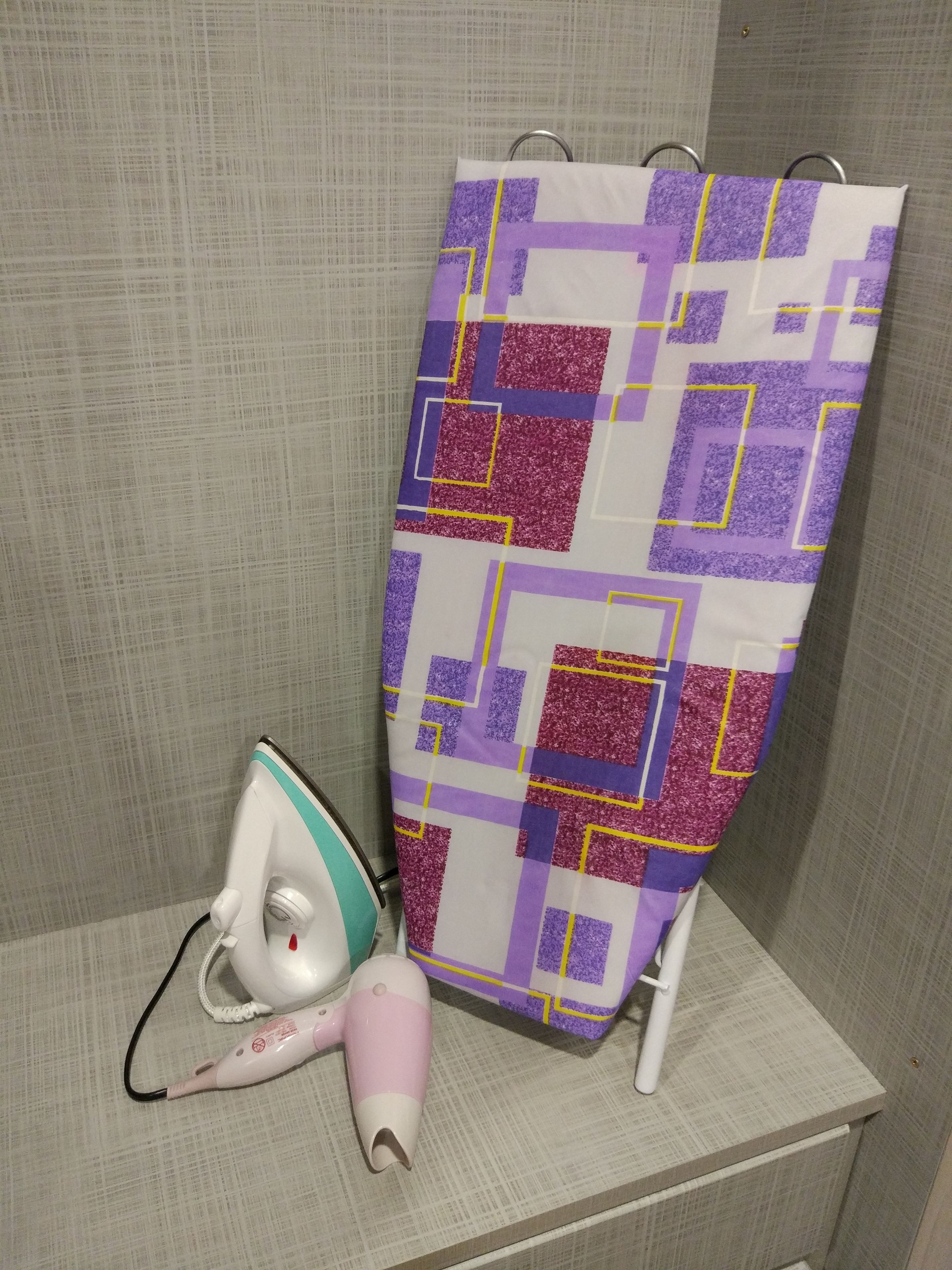 Une salle de bain avec un rideau de douche à motifs, un appareil à vapeur portatif blanc et un fer à repasser blanc posé sur une étagère.