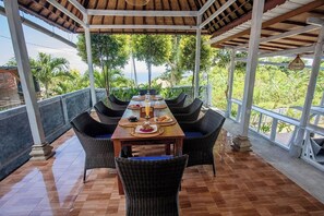 A covered outdoor dining area with a table set for two, surrounded by wicker chairs, overlooking a lush landscape. - Tatak Bunut Villa Nusa Ceningan (Ceningan Island)