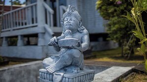 Eine stehende Steinfigur mit einer Schale auf einem Sockel, vor einem Gebäude mit einem Balkon.
