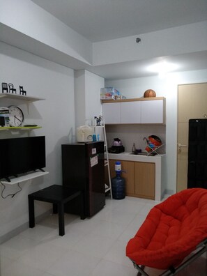 A compact living space with a television, a refrigerator, a water dispenser, a red chair, and a kitchenette area. - RJB at Ayodhya Tangerang (Tangerang)