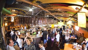 A crowded restaurant with high ceilings and exposed beams, featuring a bar area and multiple dining tables. - Nomads Hotel - Near San Clemente Pier (San Clemente)