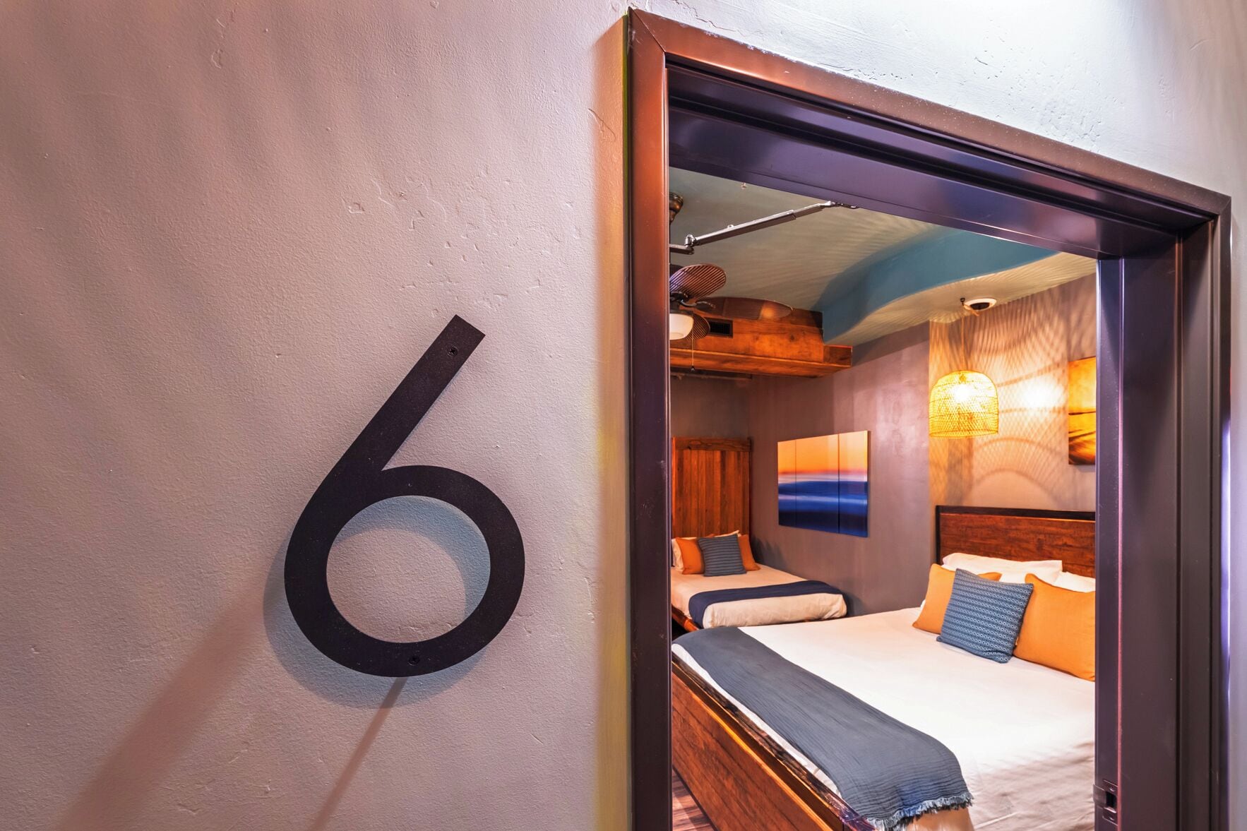 Une chambre d’hôtel avec un grand numéro « 6 » sur le mur, deux lits avec des têtes de lit en bois, un ventilateur de plafond et un tableau accroché au mur.