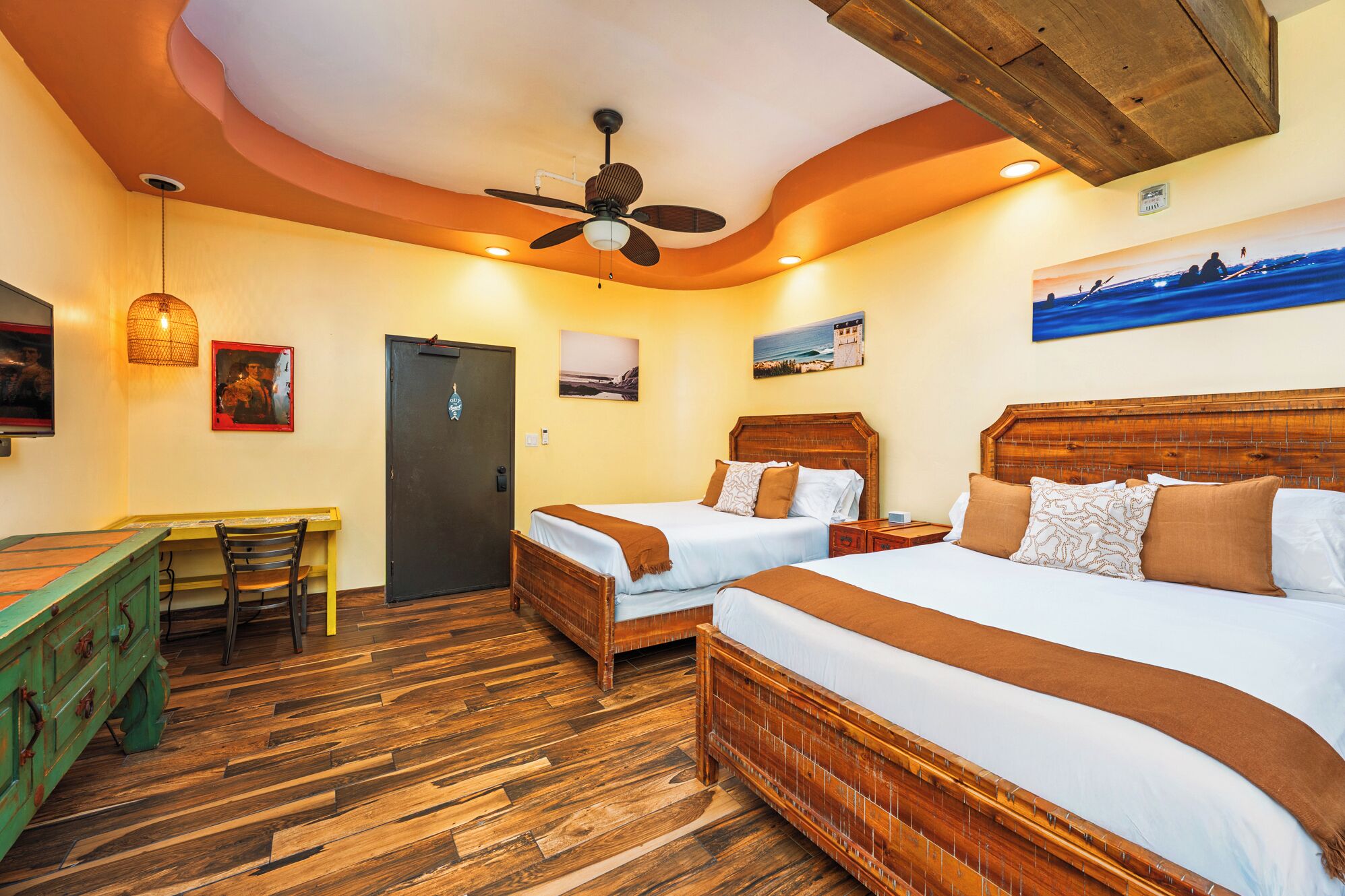 Quarto de hotel com duas camas, piso de madeira, ventilador de teto e obras de arte nas paredes.