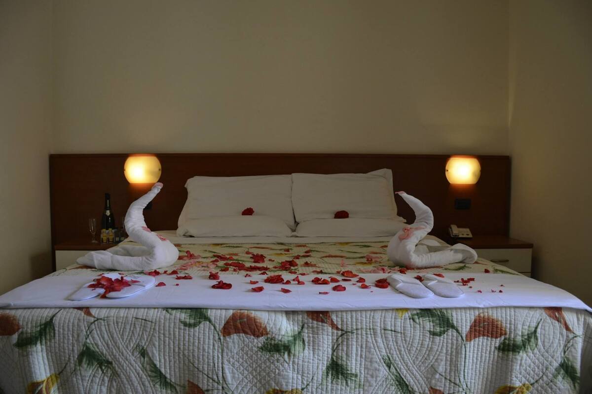 A neatly made bed with rose petals and swan-shaped towels, a bottle, and a glass on the nightstand.