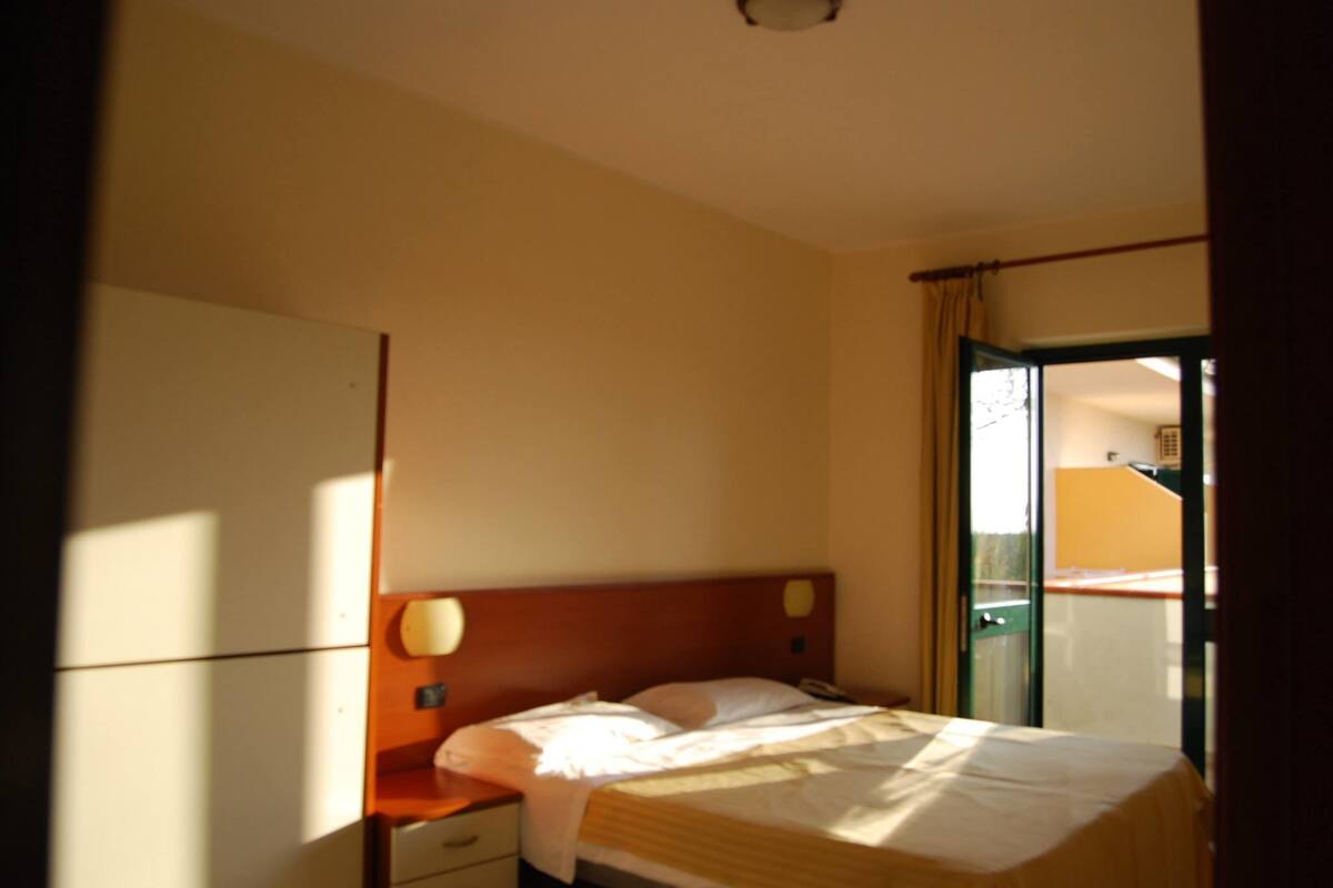 A hotel room with a bed, a nightstand, a wall-mounted lamp, and a sliding door to a balcony.