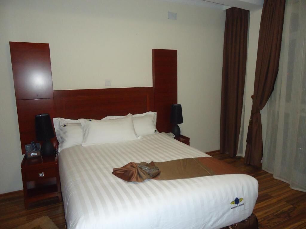 A hotel room with a large bed, wooden headboard, two bedside tables with lamps, and a window with curtains.
