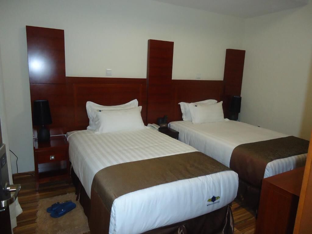 Une chambre d’hôtel avec deux lits, des têtes de lit en bois, des tables de chevet avec des lampes et un petit bureau avec une chaise.