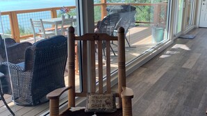 Una silla de madera con cojín y un cartel que dice 'Bienvenidos a nuestra cabaña', una terraza de madera con mobiliario exterior, y un ventanal con vista a un lago.