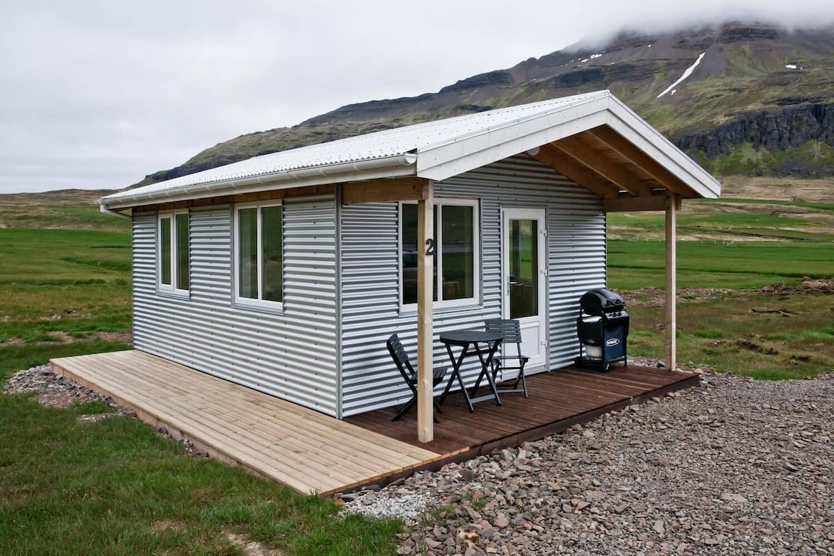 Una cabaña pequeña de un nivel con fachada de metal y una terraza de madera, que incluye una parrilla y asientos al aire libre.