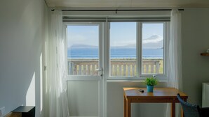 Een kamer met uitzicht op zee en een berg, een houten tafel met een plant en een stoel.