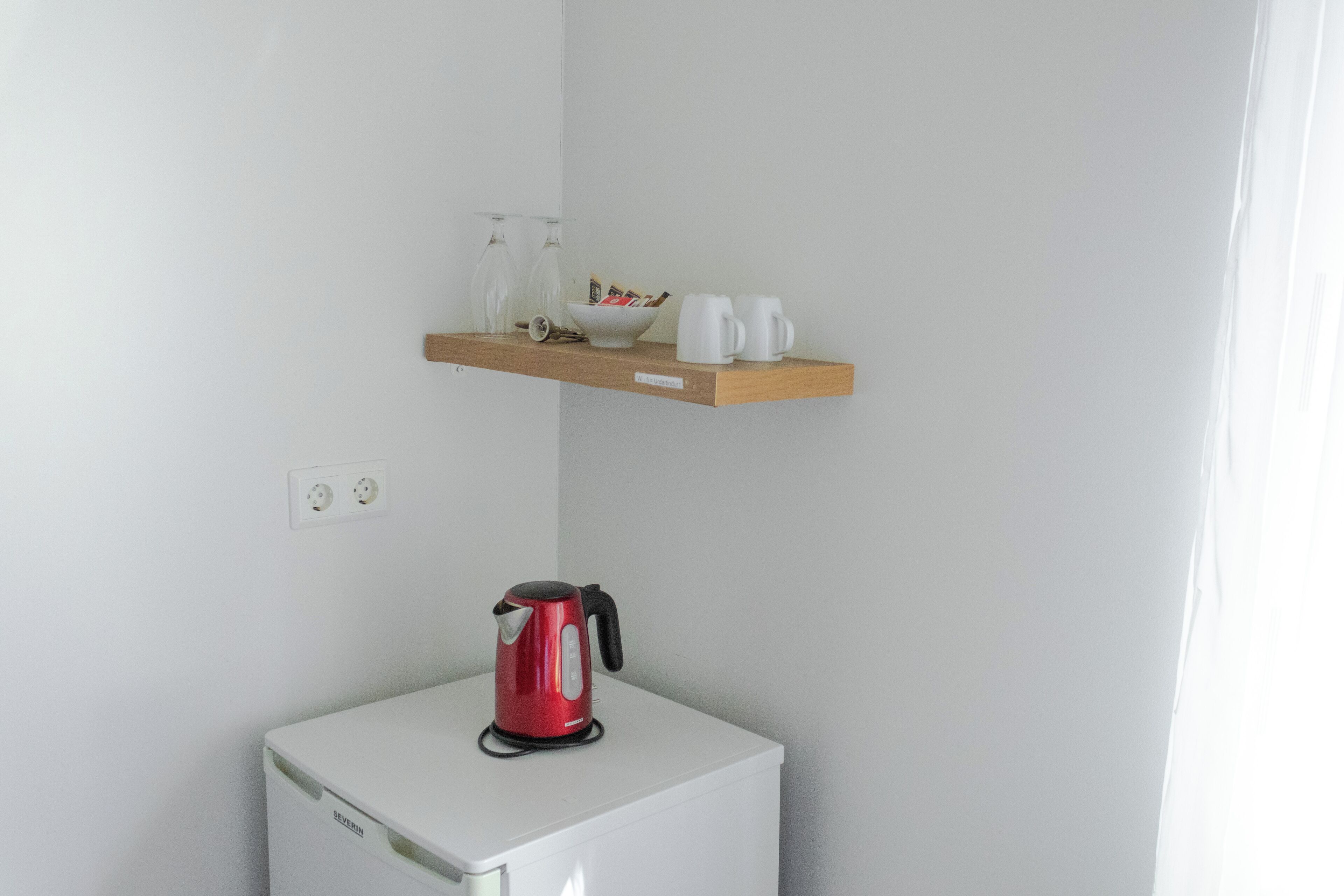 Eine kleine Küchenzeile mit einem weißen Kühlschrank, einem roten Wasserkocher und einem Holzregal darüber, auf dem sich eine Schüssel, Gläser und Tassen befinden.
