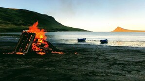 Een strand met een vuur op het zand, boten in het water en een heuvel op de achtergrond.