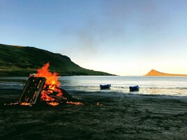 Una playa con una fogata sobre la arena, barcos en el agua y una colina al fondo.