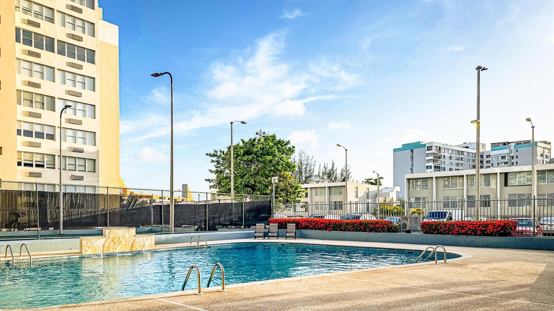A swimming pool with a clear blue water, surrounded by a fence, and adjacent to a building with multiple windows and balconies.