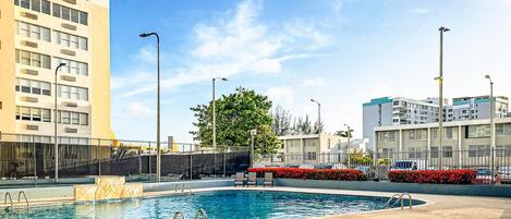 A swimming pool with a clear blue water, surrounded by a fence, and adjacent to a building with multiple windows and balconies.