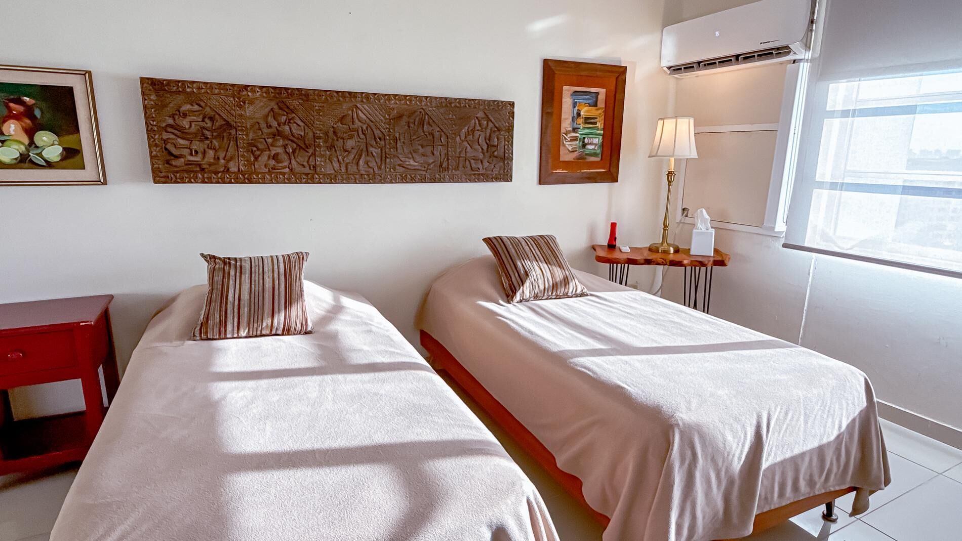 A hotel room with two beds, a red side table, a lamp, and artwork on the wall.