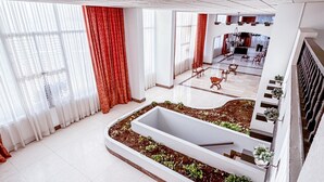 A spacious hotel lobby with a central garden area, large windows with sheer curtains, and a modern staircase.