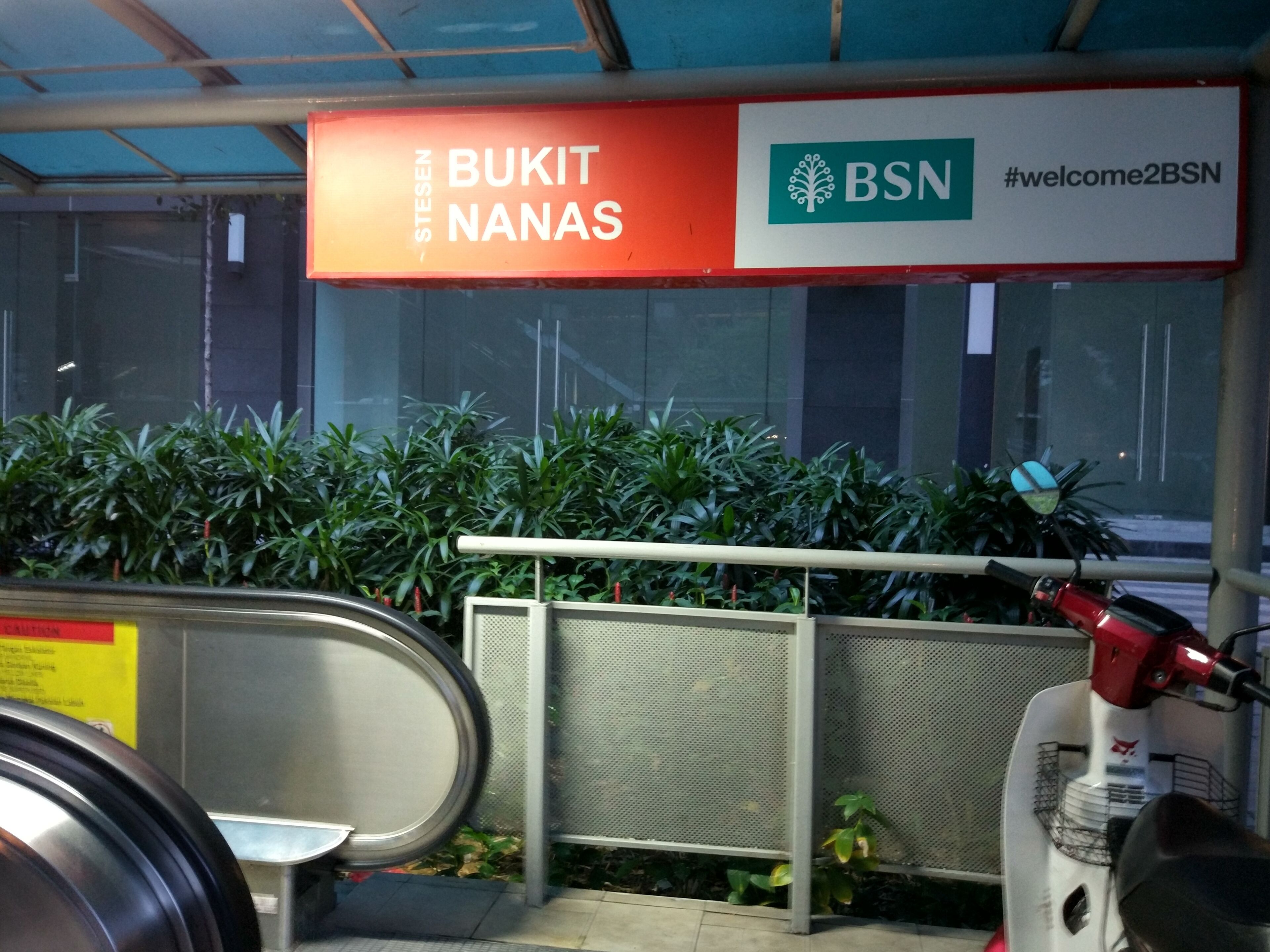A sign with 'BUKIT NANAS' and 'BSN' logos, a metal railing, and a red scooter.