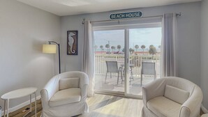 A room with two white armchairs, a small table, a lamp, and a view of the beach through a sliding glass door.