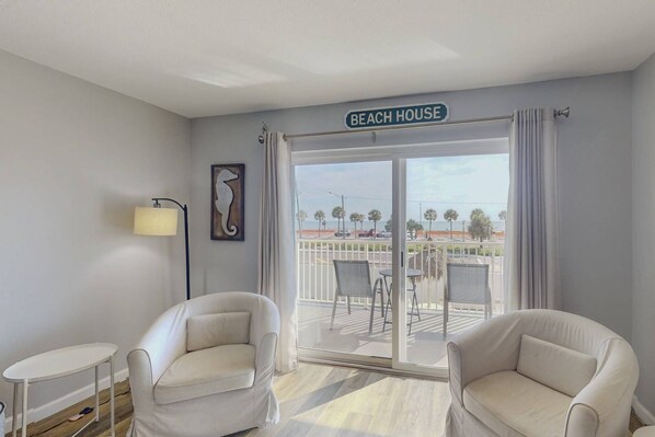 A room with two white armchairs, a small table, a lamp, and a view of the beach through a sliding glass door.