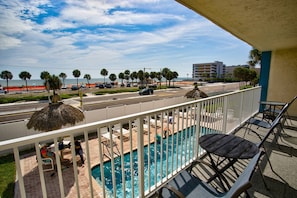 A balcony with a pool, chairs, and a view of the beach and ocean.