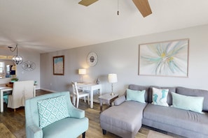 Ein Wohnzimmer mit grauem Sofa, einem türkisfarbenen Sessel, einem Essbereich mit weißem Tisch und einer Küche im Hintergrund.