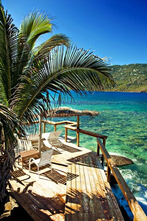 A wooden deck with lounge chairs and a thatched umbrella, overlooking clear blue water. - Casa Jo Mama so private clothing is optional!! (St. Thomas)