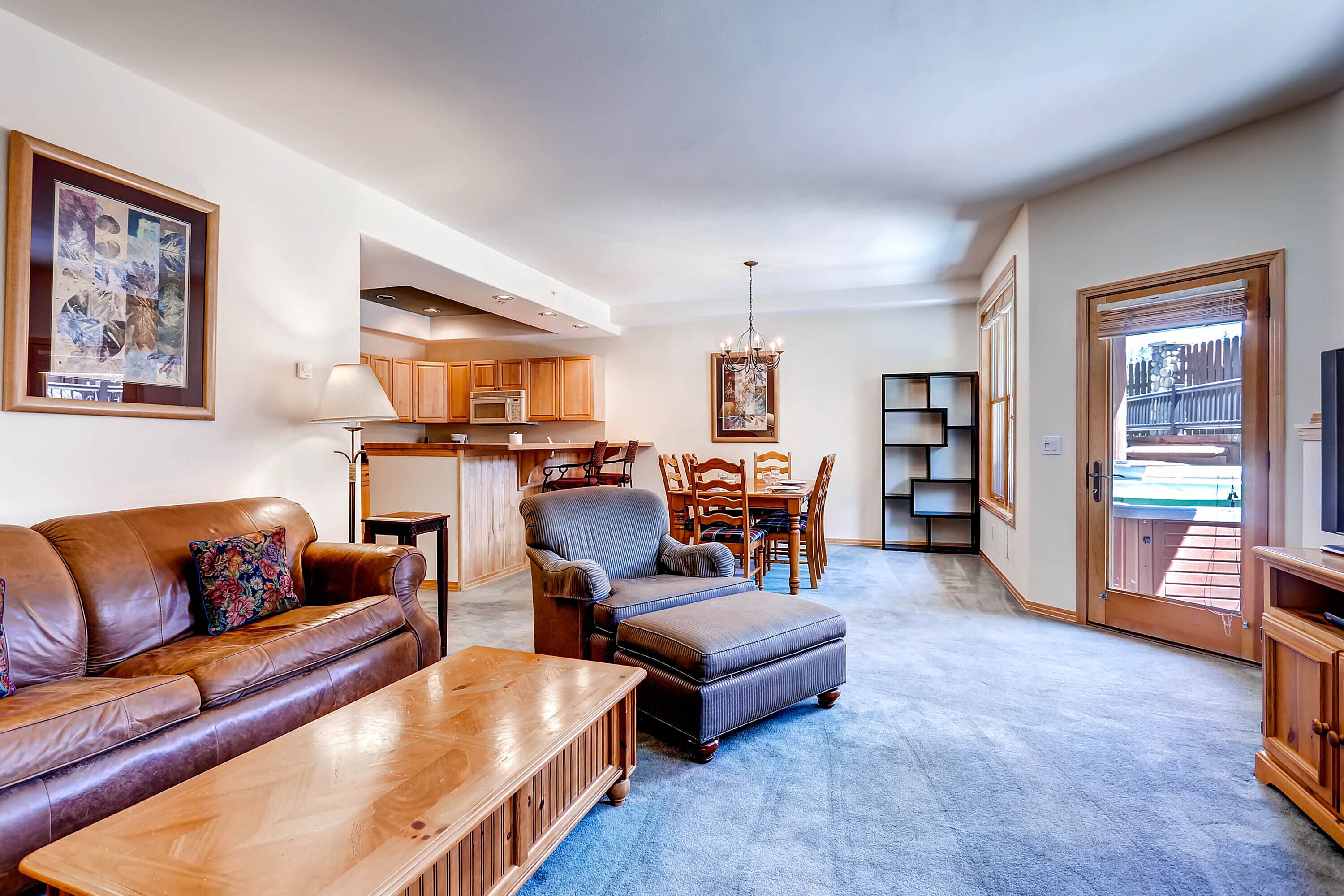 A living room with a sofa, ottoman, coffee table, and a dining area with chairs and a table. There is a kitchen in the background with cabinets and a microwave.