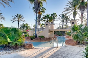 Zona de piscina con palmeras, un puente y una torre del reloj.