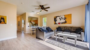 A living room with a grey sofa, a white coffee table, and a ceiling fan. There is a kitchen in the background with wooden cabinets and a dining area. - 4BR Regal Palms Townhome by FS FL VH-154 (Davenport)