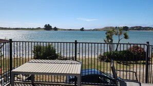 A deck with a table and chairs overlooking a lake, with a car parked nearby and a fence in the foreground. - The Pink Pipi Karitane (Karitane)