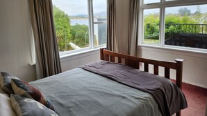 A bedroom with a bed, curtains, and a view of greenery and a body of water. - The Pink Pipi Karitane (Karitane)