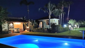 Une piscine éclairée la nuit, avec un espace patio et une maison en arrière-plan.