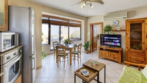 A modern kitchen with stainless steel appliances, a dining area with wooden chairs and a table, a television, and a ceiling fan.