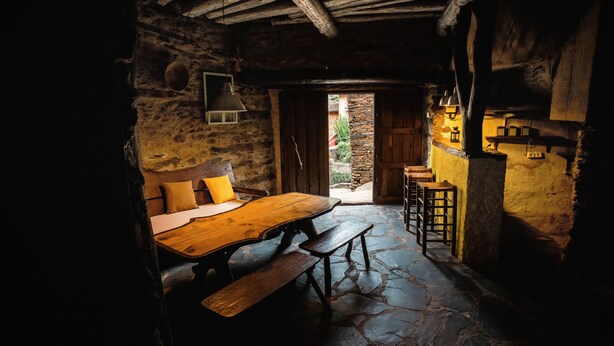 A rustic stone-floored dining area with a wooden table and benches, a stone wall with a wooden door, and a hanging light fixture. - Self catering El Mirador de Robledillo for 3 people (Robledillo de Gata)
