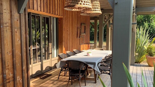 En træterrasse med et bord og stole, omgivet af store vinduer og en hængende lampe.