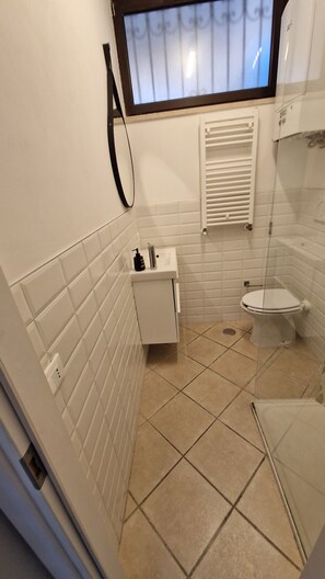 Un baño pequeño con azulejos blancos, inodoro, lavamanos y ventana con reja de metal.
