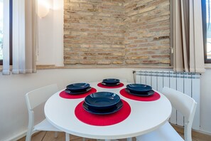 Una mesa redonda blanca con cuatro platos azules sobre caminos de mesa rojos, rodeada de sillas blancas, frente a una pared de ladrillo con una ventana y cortinas.