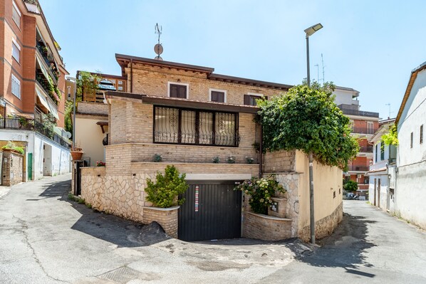 A narrow street with a building featuring a black gate, stone walls, and potted plants. - Vatican Apartment. (Roma)