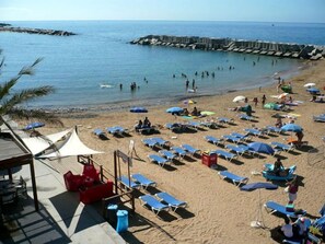 A beach with rows of sun loungers and umbrellas, a pier in the background, and a clear view of the sea.